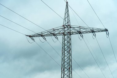 Yüksek bir elektrik direği, arka plandaki gri bulutlara karşı yükseliyor, elektrik hatları birden fazla yöne uzanıyor, serin bir günde altyapı sergileniyor..