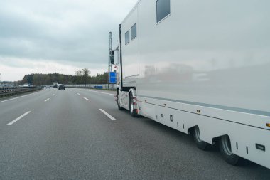 Ağaçlar ve açık alanlarla çevrili çok şeritli bir otoyolda geniş beyaz bir kamyonun sürdüğü görülüyor. Gökyüzü bulutlu, kasvetli bir günü işaret ediyor..