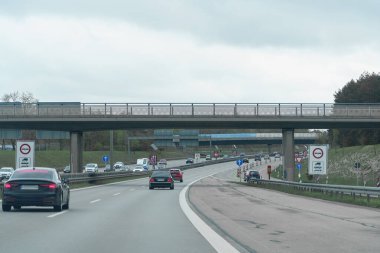 Arabalar bulutlu bir günde üst geçidin altındaki yoğun bir otoyolda yol alırlar. Yol işaretleri, asfalt yolda iki yönde giden araçlar olarak trafik kurallarını gösteriyor..