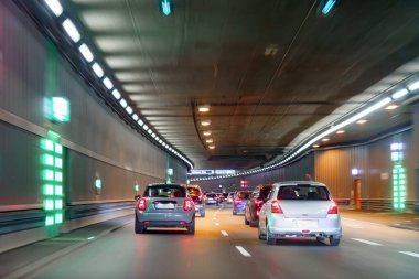 Araçlar yeşil işaretlerle kaplı parlak ışıklı bir tünelden geçiyor. Yoğun sahne, ilerideki yolu aydınlatan farlarla şehir yaşamını yakalıyor..