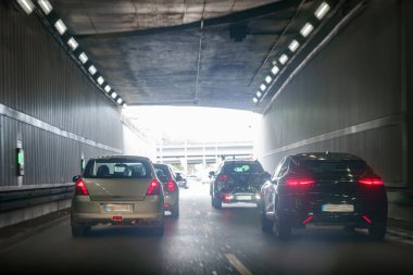Akşamları yoğun saatlerde araçlar beton duvarlar ve tepeden aydınlatma ile çevrili karanlık bir tünelden geçiyor. Arka plandaki gri fonda kırmızı ışık yanıyor..