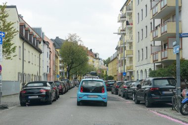Bulutlu bir gökyüzünün altında park halindeki araçlarla ve çeşitli apartmanlarla dolu bir yerleşim yeri. Ağaçlar kenar boyunca yeşillik sağlar, bu da onun kentsel bir alanda olduğunu gösterir..