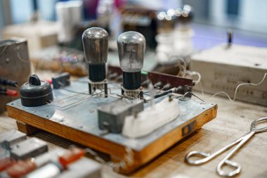 Çeşitli eski elektronik parçalar ahşap tezgah boyunca yayılır. Yakınlarda kablolar ve aletlerle, restorasyon çalışmalarını ön plana çıkaran iki vakum tüpü ön plana çıkarıldı.