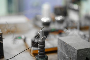 Hassas bir enstrüman atölye masasında belirgin bir şekilde duruyor. Etrafında, gündüz saatlerinde canlı bir imalat ortamının oluştuğunu gösteren çeşitli alet ve ekipmanlar var..