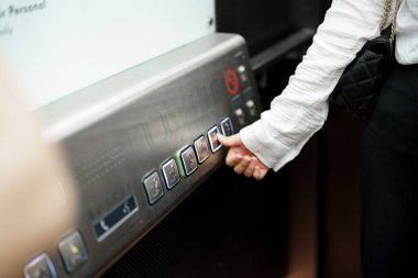 Çağdaş bir binada bir el asansör düğmesine basıyor. Ortam aydınlık, gündüz olduğunu gösteriyor. İnsanlar arka planda hareket ederken görülebilir, tipik bir şehir atmosferi..