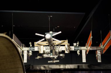 Karanlık bir müzenin tavanından sarkan uzay istasyonunun ayrıntılı bir modeli sergilenmektedir. Ziyaretçiler, insan yaratıcılığının ve uzaydaki teknolojik gelişmelerin bu temsilini gözlemliyorlar..