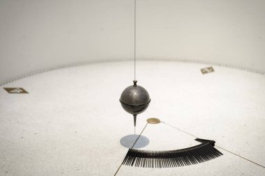 Metal bir Foucault sarkacı tavandan sarkan, güneş ışığı altındaki bir müzenin diyagramlarıyla işaretlenmiş dairesel bir tabanda sallanarak Dünya 'nın gerçek zamanlı dönüşünü gösteriyor..