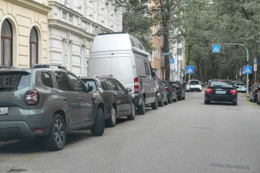 Sessiz bir caddenin her iki tarafında ağaçlarla çevrili birden fazla araç park halinde. Bu hareketli mahallede kentsel yaşamı ön plana çıkaran bir araba geçiyor..