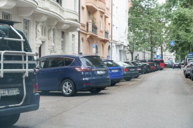 Canlı bir şehir caddesi, çeşitli modellerden park edilmiş arabalarla doludur ve tipik bir şehir atmosferi yaratır. Yakınlardaki konut binaları, yeşillik sergilenen sahneyi çerçeveliyor..