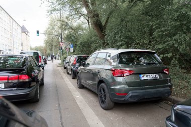Arabalar dar bir şehir caddesi boyunca yakın bir şekilde park edilmiş, kenarına bir araç yerleştirilmiş ve trafik lambası yeşil görünüyor. Bereketli yeşillik sahneye karşıtlık ekler.