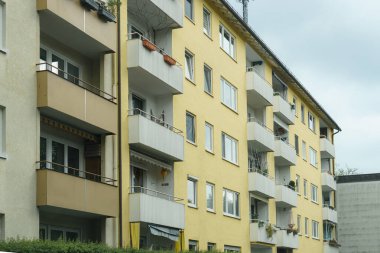 Çok katlı bir bina, canlı sarı bir dış görünüme ve bitkilerle süslenmiş sayısız balkona sahiptir. Sahne, bulutlu bir günde tipik bir şehir ortamını yakalıyor..