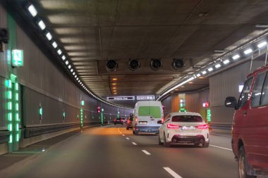 Arabalar, yaklaşan çıkışlara yaklaşırken parlak ışıklarla aydınlatılan geniş bir şehir tünelinde sorunsuzca ilerlerler. Yol açık tabela ve araçların karışımıyla dolu..