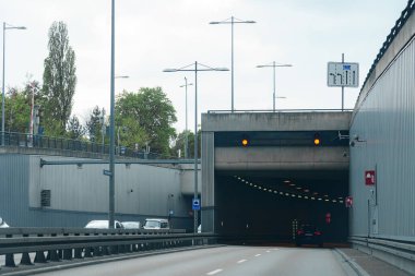 Arabalar tünele giden büyük bir yolda yol alırlar. Trafik işaretleri, yapının bulutlu gökyüzünün altında belirdiğini gösteriyor..