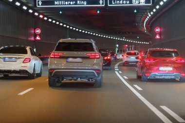 Bir grup araba gece vakti iyi aydınlatılmış bir tünelden geçiyor. Kırmızı uyarı sinyalleri ve yol işaretleri çeşitli renklerdeki parlak araçların arasında trafik akışına rehberlik ediyor.