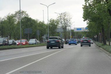 Arabalar yeşilliklerle çevrili işlek bir şehir caddesinde her iki yönde de seyahat ederler. Atmosfer bulutlu gökyüzüne rağmen sakin ve araçlar çeşitli renk ve boyutlarda..