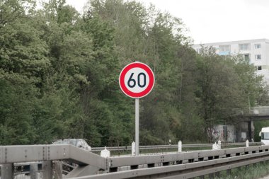 Otobanın yanında saatte 60 kilometre hız sınırı işareti var. Bölgede yemyeşil ağaçlar ve doğaya yakın kentsel bir ortamı gösteren yakınlardaki binalar yer alıyor..