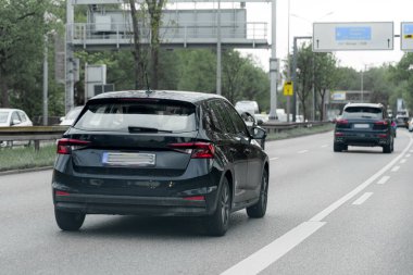 Bir siyah hatchback işlek bir şehir yolu boyunca seyahat ederken başka bir araba da onu yakından takip ediyor. Ağaçlar cadde boyunca uzanır ve bulutlu bir günde trafik işaretleri görünür..