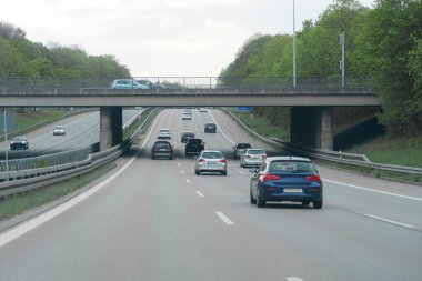 Araçlar, beton bir köprünün altındaki geniş bir otoyolda iki yöne de gidiyor. Yeşillik yol boyunca yemyeşil ve gri bulutlar gökyüzünü dolduruyor. Bu da bulutlu havayı gösteriyor..