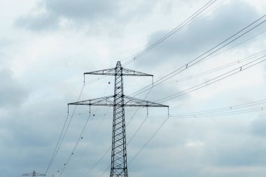 Uzun elektrik hatları, öğleden sonra aydınlanan gri bulutların zemininde belirgin bir şekilde yükseliyor. Sanayi manzarası elektrik dağıtımının altyapısını vurguluyor.