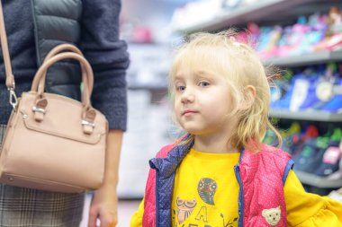 Sarı saçlı genç bir kız bir ayakkabı mağazasını keşfediyor, ilginç bir şekilde parlak renkli ayakkabılara bakıyor. Sarı bir gömlek ve yelek giyiyor, bir yetişkinle alışverişin keyfini çıkarıyor..