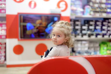 Kıvırcık saçlı genç bir kız bir oyuncakçıda parlak bir bankta oturuyor. Büyük bir ekranda renkli oyuncaklarla çevrili bir animasyon dizisini izlerken gülümseyerek arkasına bakıyor..