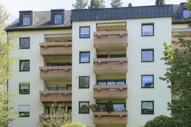 Çağdaş bir konut yapısında, bir banliyö mahallesinde yemyeşil bitkilerle çevrili birden fazla balkon bulunur. Güneş ışığı sahneyi aydınlatır..