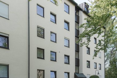 Birden fazla penceresi olan çok katlı bir apartman yemyeşil ağaçlarla çevrilidir. Sahne, sakin bir banliyö mahallesinin ön cephesini aydınlatan parlak güneş ışığını yakalıyor..
