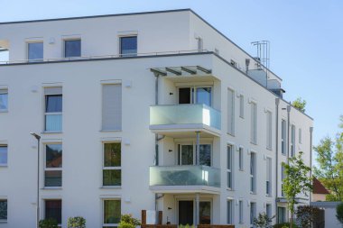 Çağdaş apartmanda birden fazla balkon ve geniş pencereler bulunmaktadır. Tasarım yeşilliği içeriyor ve açık gökyüzünün altında bir şehir merkezinde yer alıyor..