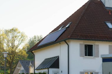 Kırmızı kiremitli çatısında güneş panelleri olan modern bir ev yumuşak sabah güneşiyle yıkanır. Arka planda yemyeşil ağaçlar ve diğer evler görülebiliyor ve huzurlu bir ortamı vurguluyorlar..