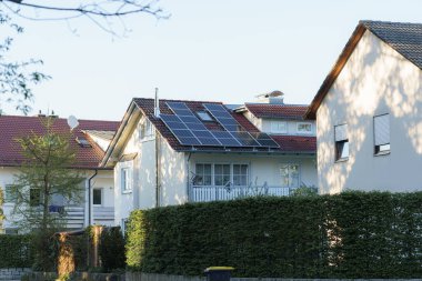 Güneş panelleriyle donatılmış rahat bir ev komşu evin yanında oturur. Ilık sabah güneşi yeşil çitlerin üzerine yumuşak gölgeler düşürüyor ve sakin bir atmosfer yaratıyor..