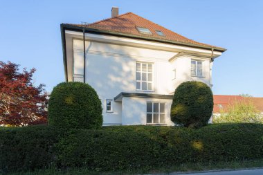 Beyaz bir ev, parlak mavi bir gökyüzünün altında düzgünce budanmış çitlerin arasında dimdik duruyor. Mimaride büyük pencereler ve eğimli bir çatı var. Huzurlu bir ortama yerleştirilmiş..