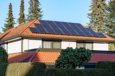 Modern bir evin çatısındaki güneş panelleri enerji için güneş ışığını yakalıyor. Kurulumda uzun ağaçlar ve sürdürülebilir canlıları gösteren açık mavi bir gökyüzü yer alıyor..