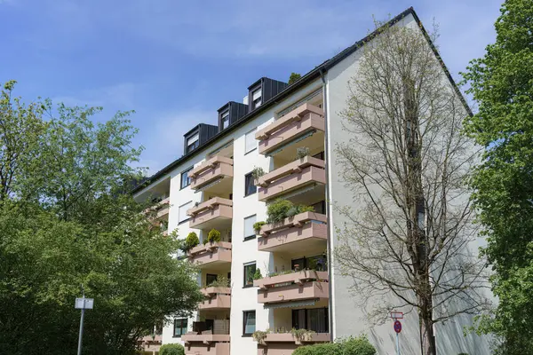 Modern bir konut binasında, berrak mavi gökyüzü ve yeşilliğin altında huzurlu bir mahallede bulunan bitkilerle süslenmiş çok sayıda balkon bulunuyor..