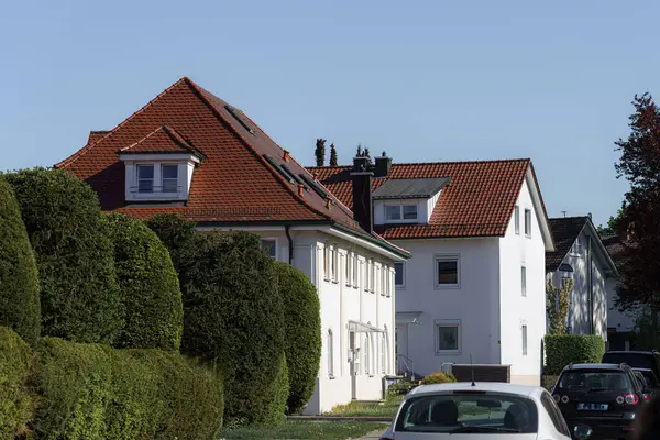 Yerleşim yerlerinin manzaralı bir manzarasında, yemyeşil ile çevrili kırmızı çatıları olan iyi bakımlı evler yer alır. Güneş ışığı sonbahar atmosferini güçlendirir, rahat bir atmosfer yaratır..