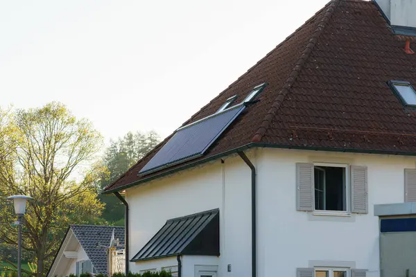 Kırmızı kiremitli çatısında güneş panelleri olan modern bir ev yumuşak sabah güneşiyle yıkanır. Arka planda yemyeşil ağaçlar ve diğer evler görülebiliyor ve huzurlu bir ortamı vurguluyorlar..