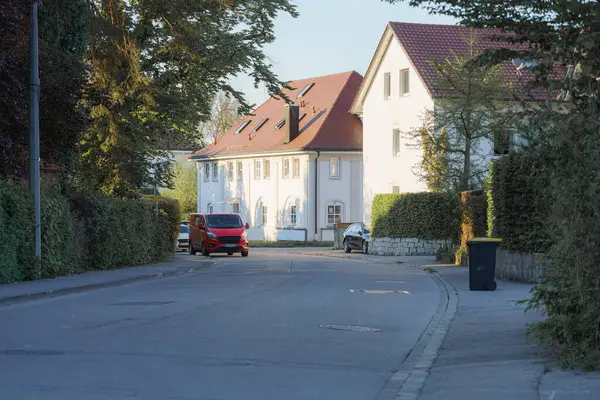 Güneşli bir öğleden sonra yeşil ve konut binalarıyla çevrili sakin bir caddede kırmızı bir araç park halindedir. Bölge huzurlu bir banliyö yaşam tarzını yansıtıyor..