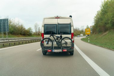 Arkasına güvenli bir şekilde monte edilmiş bir dağ bisikletiyle otoyolda giden beyaz bir minibüs. Ağaçlar çiçek açıyor, baharın gelişini sergiliyorlar. Yol bizi uzaklara götürür..