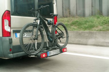 Siyah bir bisiklet bir otoyol boyunca seyahat ederken güvenli bir şekilde bir minibüsün arkasına bağlanır. Arka plandaki yeşillik, aydınlık bir günde manzaralı bir yol izlenimi veriyor. Açık hava aktiviteleri için mükemmel..