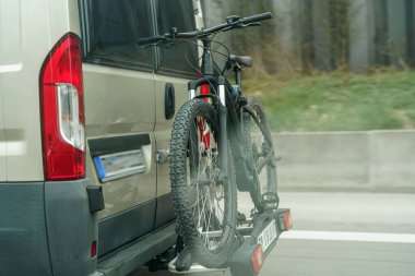 Bir minibüs otobanda gidiyor. Arka taraftaki askıya monte edilmiş iki bisiklet var. Kırsal bölgede açık bir gökyüzünün altında yeşil çimenler ve ağaçlar var..