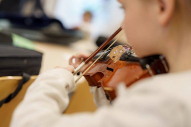 Genç bir çocuk sıcak ve davetkar bir müzik sınıfında keman çalmaya dikkatle odaklanır. Çantalar ve enstrümanlarla çevrili atmosfer öğrenmek için teşvik edici..