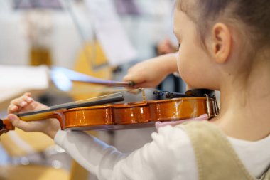 Genç bir kız diğer öğrencilerle çevrili bir müzik sınıfında keman tekniğine odaklanır. Yumuşak doğal ışık, neşeli sınıf atmosferini doldurur..