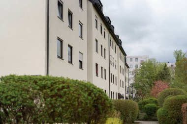 Çağdaş bir yerleşim yeri kentsel alandaki gür çalıların ve ağaçların arasında duruyor. Bulutlu gökyüzü mekanın sakin atmosferini arttırıyor. Bu da huzurlu bir yaşam ortamı olduğunu gösteriyor..