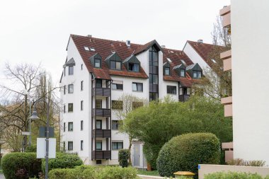 Modern bir konut binasında çok sayıda balkon ve yemyeşil alanlar arasında çeşitli şekiller yer alır. Atmosfer bulutlu bir günde kentsel ama sakin bir banliyö ortamını yansıtıyor..