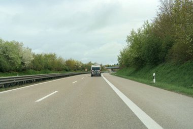 Bir araç ağaçlar ve çalılarla kaplı ıssız bir otoyolda ilerliyor. Bulutlu gökyüzü sakin bir atmosfer yaratarak sakin bir sürüş deneyimi yaratıyor..