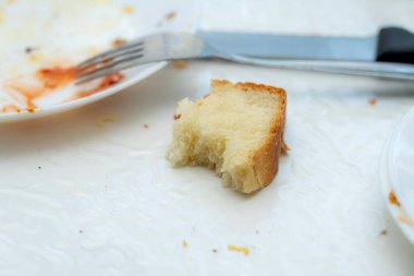 Bir yemeğin kalıntıları yanında çatal ve bıçakla tabakta duran küçük bir parça ekmek içerir. Aydınlık ve davetkar bir ortam, sıradan bir yemek atmosferini yansıtıyor..