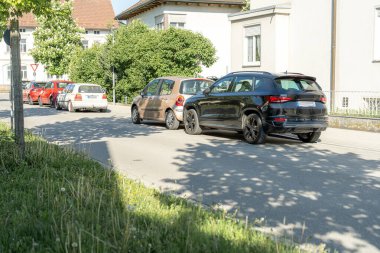 Huzurlu bir yerleşim yolunda, birkaç küçük arabanın yanına siyah bir SUV park edilmiştir. Sahne güneşli bir öğleden sonra yeşilliğin yakınında sakin bir atmosfer yaratıyor..