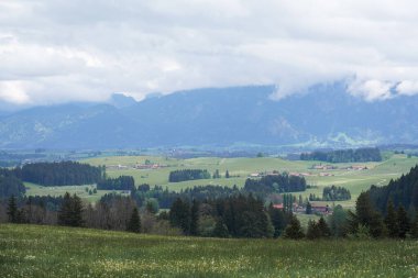 Uçsuz bucaksız manzaralar, çiftlikler ve evler ile benekli yeşil tarlaları besler. Uzak dağlar bulutların altında belirir, baharda huzurlu ve resimli bir manzara yaratır..