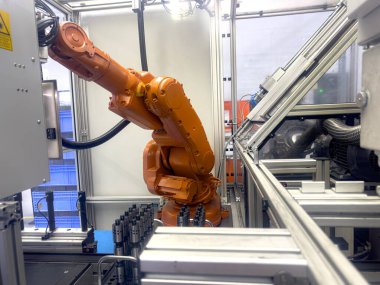Turuncu kaplamalı bir endüstriyel robot kolu modern bir üretim tesisinde özenle çalışıyor. Çevre gelişmiş makine ve ekipmanlarla dolu..