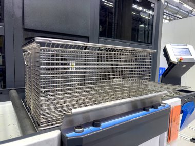 İyi ışıklandırılmış bir endüstriyel tesisteki bir kontrol istasyonunda metal bir sepet konveyör sistemi üzerinde duruyor. Çevrede modern makineler ve temiz bir çalışma alanı sergileniyor.