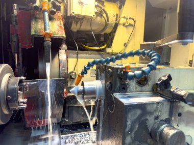 Bir makinist atölyede torna işletiyor, metal parçalarını kayganlaştırıcı ile şekillendiriyor, makine etrafında metal talaşı biriktirirken,.